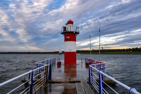 Red and white lighthouse houseboat at Houseboat am Bärwalder See, surrounded by water and a cloudy sky.