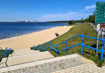 Blick auf einen Sandstrand am Bärwalder See in Deutschland mit blauem Geländer und Badenden im Wasser.