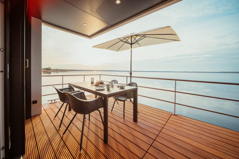 Terraza de casa flotante en Gräbendorfer See con mesa de comedor, sillas, sombrilla y vistas al lago.