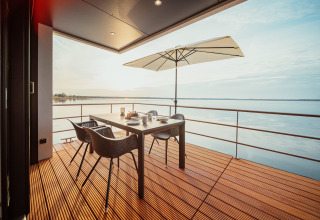 Terraza de casa flotante en Gräbendorfer See con mesa de comedor, sillas, sombrilla y vistas al lago.