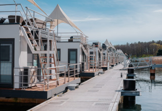 Rij moderne woonboten langs een steiger op Gräbendorfer See met kalm water en bos op de achtergrond.