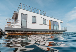 Modern husbåd ved Gräbendorfer See med hvid facade, store vinduer og rækværk, flydende på vandet.