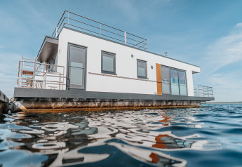 Casa flotante moderna en el Gräbendorfer See con fachada blanca, barandillas y grandes ventanas sobre el agua.