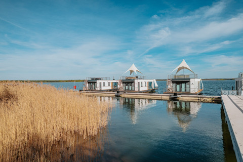 Husbåde ved Houseboat am Gräbendorfer See under blå himmel, omgivet af siv og roligt vand.
