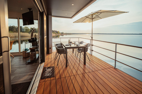 Terraza moderna de casa flotante en Gräbendorfer See con mesa, sillas, sombrilla y vista al lago.