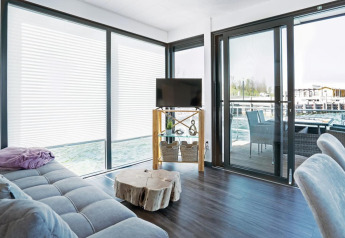 Helles Wohnzimmer auf dem Hausboot Floating 33 - LP 16, Deutschland, mit großen Fenstern und Seeblick.