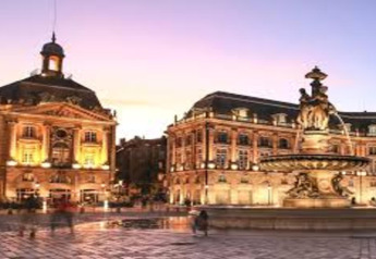 Beleuchtete historische Gebäude und ein Brunnen bei Sonnenuntergang in einer europäischen Stadt, vom Wasser aus gesehen.