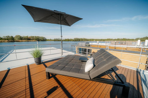 Sunny rooftop deck on a houseboat in Zehdenick with a modern lounger, umbrella, pillow, and water views.