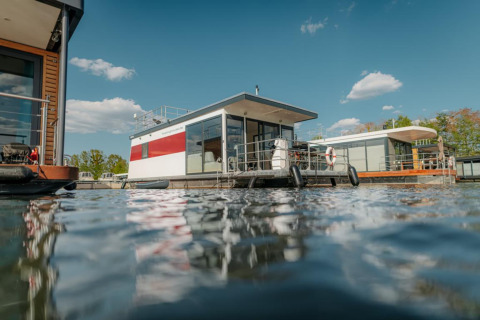 Casa galleggiante moderna a Zehdenick sull’acqua, circondata da altre case barca e cielo azzurro.