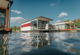 Modern houseboat in Zehdenick floating on water, surrounded by other houseboats under a blue sky.