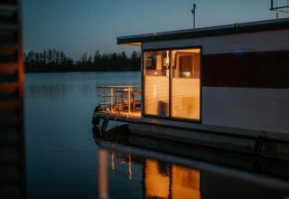Illuminated houseboat on calm water at dusk in Zehdenick, showing reflections and distant trees.
