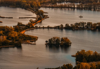 Luftfoto af sølandskab med øer og en husbåd i Zehdenick, omgivet af efterårsfarvet skov og vand.