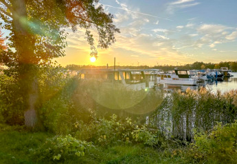 Zonsondergang aan een woonboot in Zehdenick, met bomen, riet en een rustige waterrand op de achtergrond.