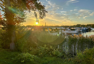 Tramonto su una houseboat a Zehdenick con alberi e canne in primo piano e acqua calma sullo sfondo.
