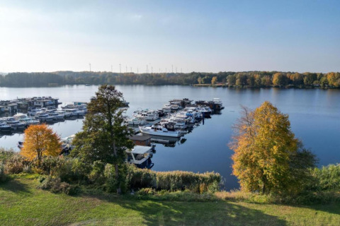 View of a marina with houseboats and boats in Zehdenick, surrounded by autumn-colored trees and calm water.