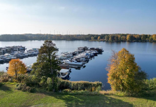 View of a marina with houseboats and boats in Zehdenick, surrounded by autumn-colored trees and calm water.