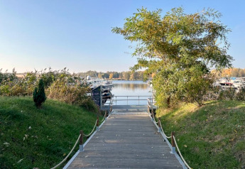 Ein Holzweg führt zu Hausbooten am Wasser in Zehdenick, umgeben von herbstlichen Bäumen und Sträuchern.