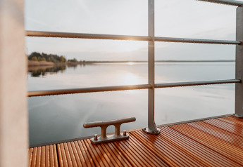 Vue depuis Houseboat am Gräbendorfer See avec terrasse en bois, rambarde et soleil reflétant sur le lac.