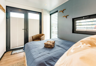 Dormitorio en una casa flotante en Zehdenick con cama doble, colcha azul y ventanas modernas.