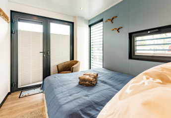Dormitorio en una casa flotante en Zehdenick con cama doble, colcha azul y ventanas modernas.