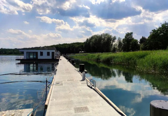 Ein modernes Hausboot am Gräbendorfer See, angedockt an einem langen Steg, umgeben von Schilf und Wald.