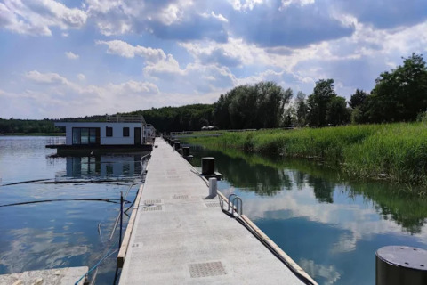 Casa flotante moderna en Gräbendorfer See, anclada a un muelle largo rodeado de juncos y árboles verdes.