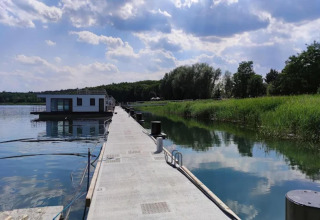 Houseboat moderno sul Gräbendorfer See, ormeggiato a un lungo molo in cemento tra canneti e alberi.
