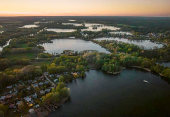 Luftaufnahme eines Hausboots am See in Zehdenick, Deutschland, umgeben von grüner Natur bei Sonnenuntergang.