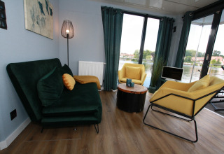 Moderne stue på husbåd 'Friedrich' i Tyskland med grøn sofa, gule stole og udsigt over vandet.
