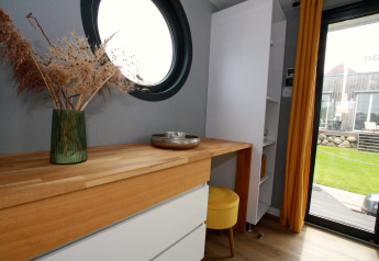 Modern interior of Houseboat Friedrich with round window, wooden desk, yellow stool and garden view.