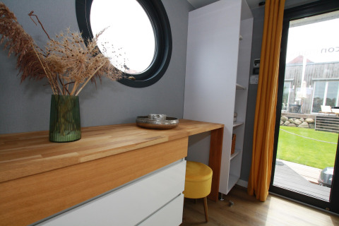 Modern interior of Houseboat Friedrich with round window, wooden desk, yellow stool and garden view.