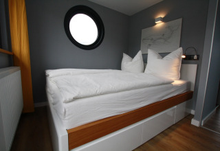 Chambre moderne sur Houseboat Friedrich avec hublot rond, murs gris et lit blanc avec bordure en bois.