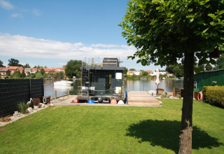 Prato verde e albero vicino alla Houseboat Friedrich ancorata su un fiume tranquillo davanti a un villaggio.