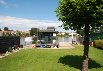 Groen gazon en boom bij de Houseboat Friedrich aan een rustige rivier met dorpszicht op de achtergrond.