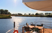 Zicht vanaf een woonboot met terras, eettafel en stoelen over een rustige rivier op een zonnige dag.