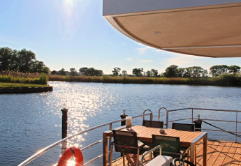 Zicht vanaf een woonboot met terras, eettafel en stoelen over een rustige rivier op een zonnige dag.