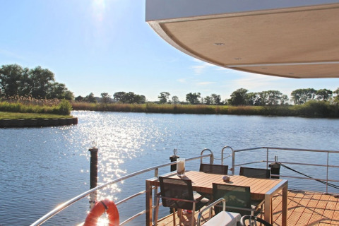 Vista desde una casa flotante con terraza, mesa de comedor y sillas hacia un río tranquilo y soleado.