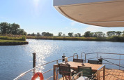 Zicht vanaf een woonboot met terras, eettafel en stoelen over een rustige rivier op een zonnige dag.