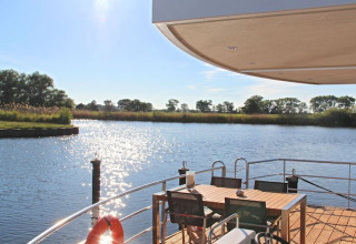 Vista desde una casa flotante con terraza, mesa de comedor y sillas hacia un río tranquilo y soleado.