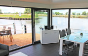 Salón moderno en casa flotante con grandes ventanales, vistas al agua y terraza con mesa exterior.