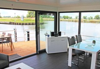 Moderne husbåd stue med store vinduer, udsigt over vandet og terrasse med udendørs spisebord.