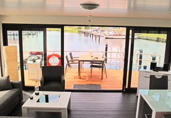 Woonkamer op een woonboot met zicht op een buitendek, haven en boten door grote schuiframen.
