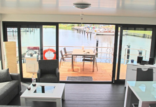Soggiorno su una houseboat con vista su un ponte esterno, il porto e barche tramite ampie porte a vetri.