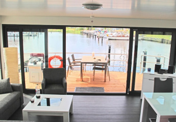 Woonkamer op een woonboot met zicht op een buitendek, haven en boten door grote schuiframen.