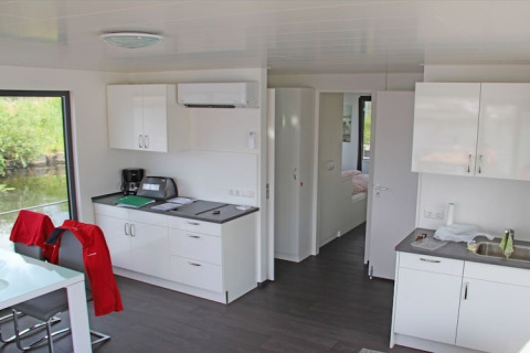 Interno di houseboat moderno con cucina bianca, tavolo da pranzo e vista sull’acqua da grandi finestre.