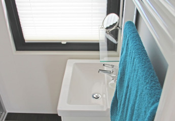 Salle de bain compacte sur une péniche avec lavabo blanc, serviette bleue et fenêtre à store vénitien.