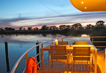 Avondsfeer op een woonboot met buitentafel, kalm water en een schitterende zonsondergang op de achtergrond.