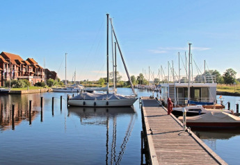 Foto von einem Hausboot und Segelbooten an einem ruhigen Hafen mit Holzsteg und Gebäuden im Hintergrund.