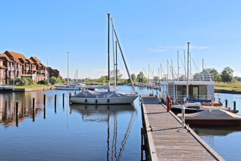 Photo of a houseboat and sailboats docked at a calm marina with wooden pier and apartments in the background.