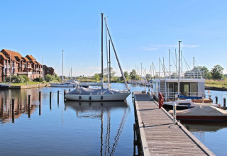 Photo of a houseboat and sailboats docked at a calm marina with wooden pier and apartments in the background.